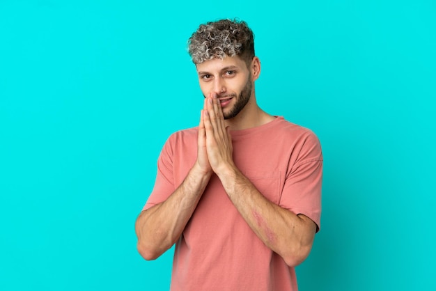 Jeune bel homme caucasien isolé sur fond bleu maintient la paume ensemble. La personne demande quelque chose