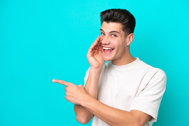 Jeune bel homme caucasien isolé sur fond bleu pointant vers le côté pour présenter un produit et chuchotant quelque chose