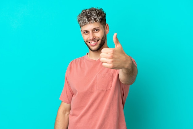 Jeune bel homme caucasien isolé sur fond bleu avec les pouces vers le haut parce que quelque chose de bien s'est produit