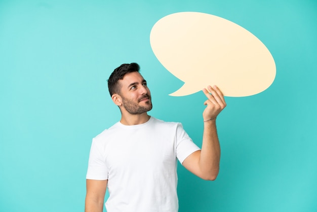 Jeune bel homme caucasien isolé sur fond bleu tenant une bulle de dialogue vide