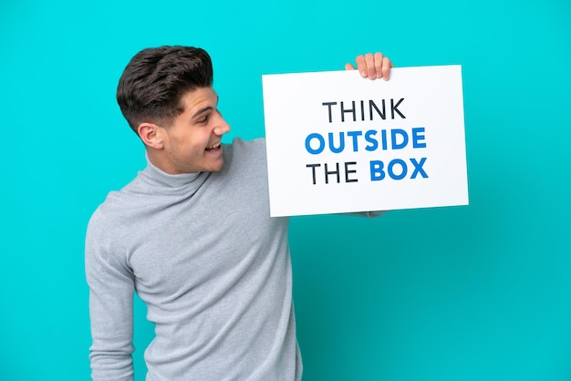 Jeune bel homme caucasien isolé sur fond bleu tenant une pancarte avec du texte Think Outside The Box