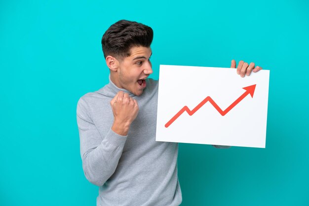 Jeune bel homme caucasien isolé sur fond bleu tenant une pancarte avec un symbole de flèche de statistiques croissante et célébrant une victoire