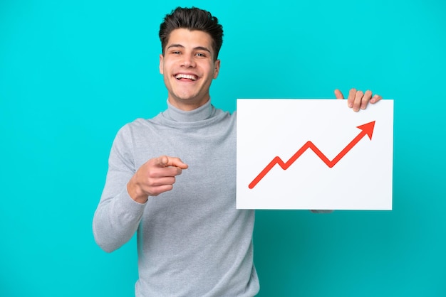 Jeune bel homme caucasien isolé sur fond bleu tenant une pancarte avec un symbole de flèche de statistiques croissante et pointant vers l'avant