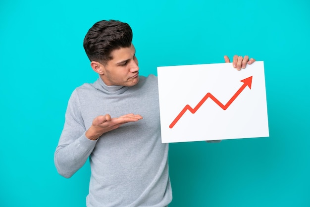 Jeune bel homme caucasien isolé sur fond bleu tenant une pancarte avec un symbole de flèche de statistiques croissante et le pointant