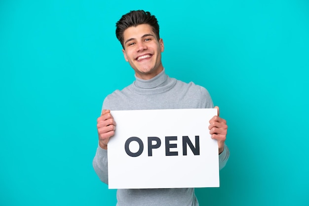 Jeune bel homme caucasien isolé sur fond bleu tenant une pancarte avec texte OUVERT avec une expression heureuse