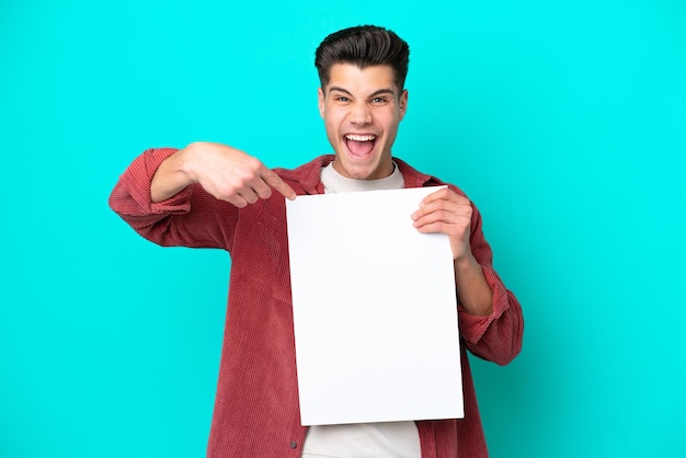 Jeune bel homme caucasien isolé sur fond bleu tenant une pancarte vide avec une expression heureuse et la pointant