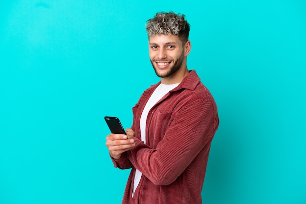 Jeune bel homme caucasien isolé sur fond bleu tenant un téléphone portable et les bras croisés