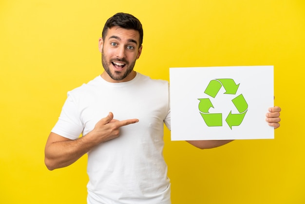 Jeune bel homme caucasien isolé sur fond jaune tenant une pancarte avec l'icône de recyclage et le pointant