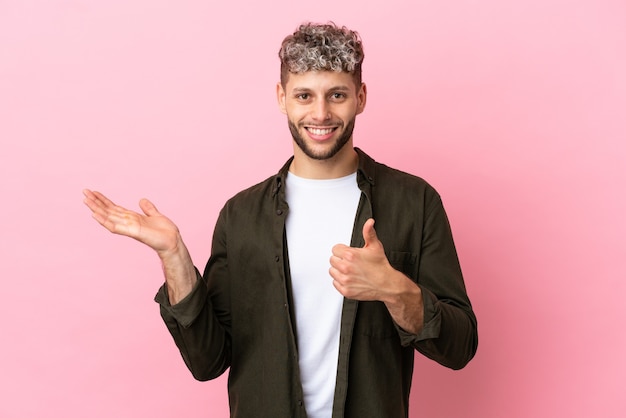 Jeune bel homme caucasien isolé sur fond rose tenant un espace de copie imaginaire sur la paume pour insérer une annonce et avec le pouce levé