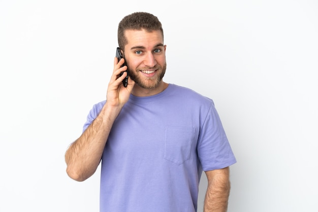 Jeune bel homme caucasien isolé sur un mur blanc en gardant une conversation avec le téléphone mobile avec quelqu'un