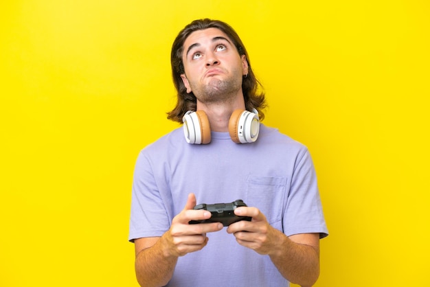Jeune bel homme caucasien jouant avec un contrôleur de jeu vidéo sur isolé sur fond jaune et levant les yeux