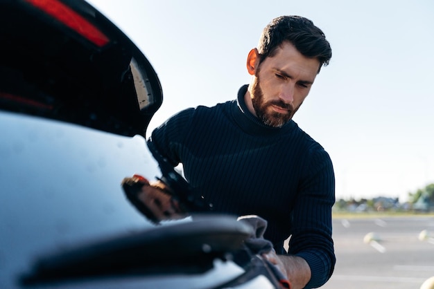 Jeune bel homme caucasien lavant et essuyant sa voiture noire moderne en plein air Homme barbu avec une lingette en microfibre grise polissant sa nouvelle voiture électrique Concept de lavage de voiture