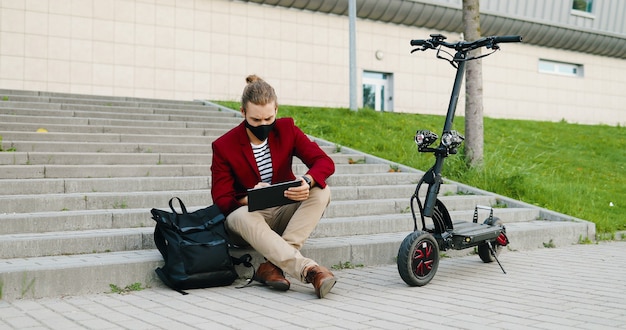 Jeune bel homme caucasien en masque et veste rouge assis sur les marches à l'extérieur, travaillant sur ordinateur gadget et navigation en ligne. Mec élégant tapant et faisant défiler sur la tablette. Scooter électrique.