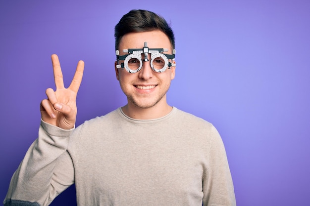 Jeune bel homme caucasien portant des lunettes optométriques sur fond violet souriant avec un visage heureux faisant un clin d'œil à la caméra faisant le signe de la victoire numéro deux