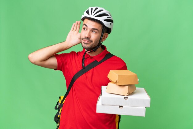 Jeune bel homme caucasien avec sac à dos thermique et tenant des plats à emporter sur fond isolé écoutant quelque chose en mettant la main sur l'oreille