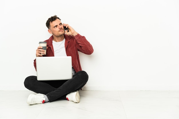 Jeune bel homme caucasien sit-in sur le sol avec ordinateur portable tenant du café à emporter et un mobile