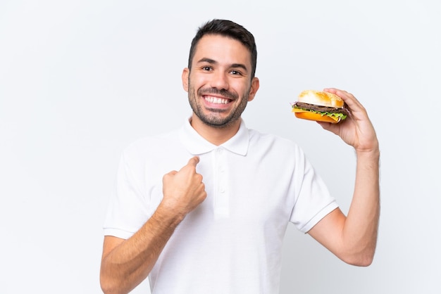 Jeune bel homme caucasien tenant un hamburger sur fond isolé avec une expression faciale surprise