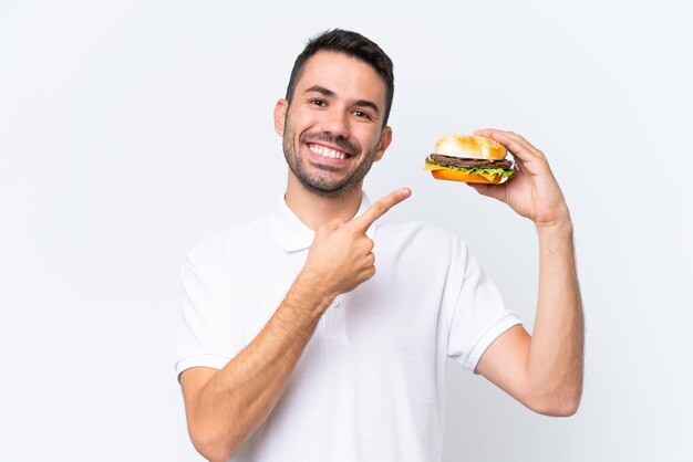 Jeune bel homme caucasien tenant un hamburger sur fond isolé et le pointant