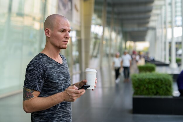 Jeune bel homme chauve tenant une tasse de café en papier et un téléphone mobile