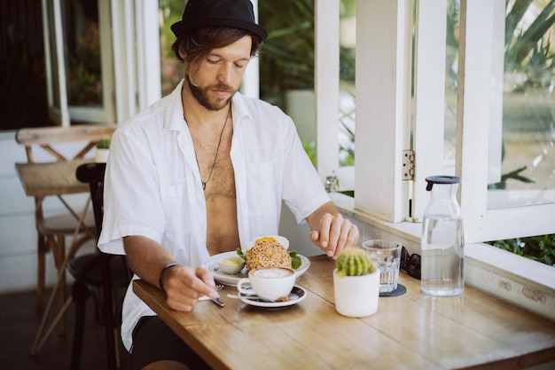 Jeune bel homme en chemise blanche ouverte, prenant son petit déjeuner dans un café avec un hamburger végétarien, buvant du café, mode de vie dans une île tropicale, vie à Bali.