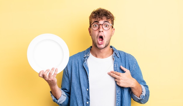 Jeune bel homme choqué et surpris avec la bouche grande ouverte, pointant vers lui-même. concept de plat vide