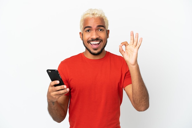 Jeune bel homme colombien isolé sur fond blanc à l'aide d'un téléphone portable et faisant signe OK