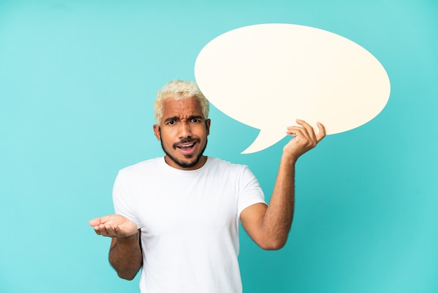 Jeune bel homme colombien isolé sur fond bleu tenant une bulle de dialogue vide avec une expression surprise