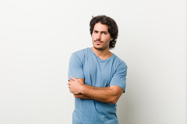 Jeune bel homme contre un mur blanc, fronçant les sourcils de mécontentement, garde les bras croisés.