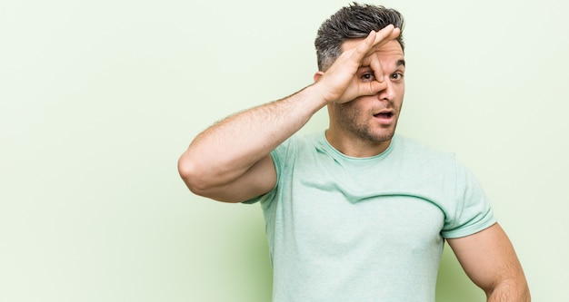 Jeune bel homme contre un mur vert excité en gardant le geste ok sur les yeux.