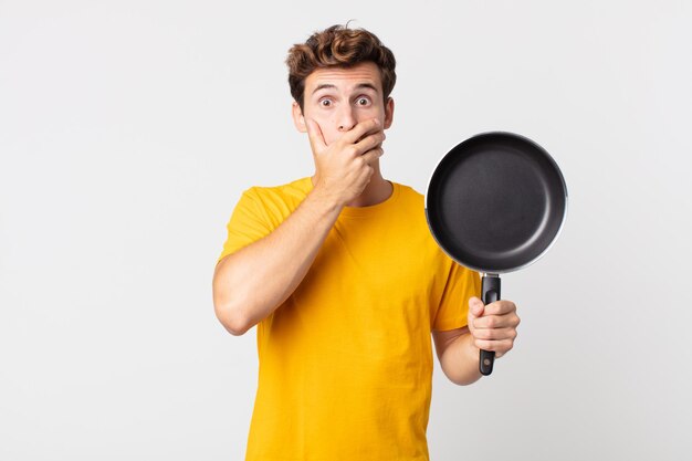 Jeune bel homme couvrant la bouche avec les mains avec un choqué et tenant une casserole