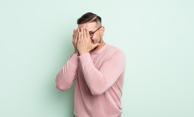 Jeune bel homme couvrant le visage avec les mains, regardant entre les doigts avec une expression surprise et regardant sur le côté