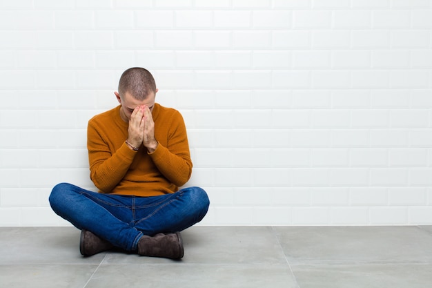 Jeune bel homme couvrant les yeux avec les mains avec un regard triste et frustré de désespoir, pleurer, vue latérale assis sur le sol