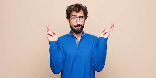 Jeune bel homme croisant les doigts avec anxiété et espérant avoir de la chance avec un regard inquiet sur un mur plat