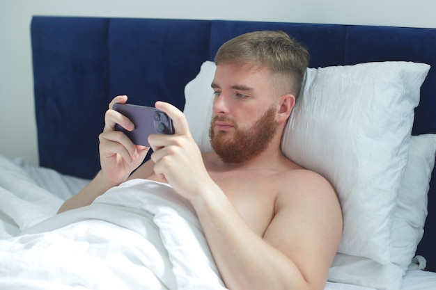 Jeune bel homme dans le lit dans la chambre à la maison le matin allongé sous une couverture blanche à l'aide d'un téléphone portable en regardant l'écran du smartphone