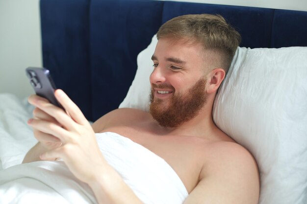 Jeune bel homme dans le lit dans la chambre à la maison le matin allongé sous une couverture blanche à l'aide d'un téléphone portable en regardant l'écran du smartphone