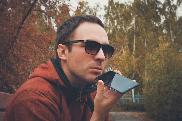 Jeune bel homme debout dans un parc en été, utilisant un téléphone portable pour écouter un message vocal ou audio enregistré. le gars parle dans le microphone pour enregistrer un message vocal. Enregistrement sur dictaphone.