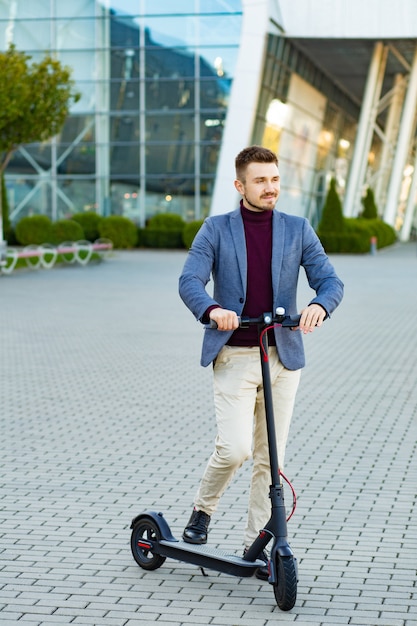 Jeune bel homme élégant avec e-scooter debout sur le trottoir près du centre d'affaires sur le coucher du soleil