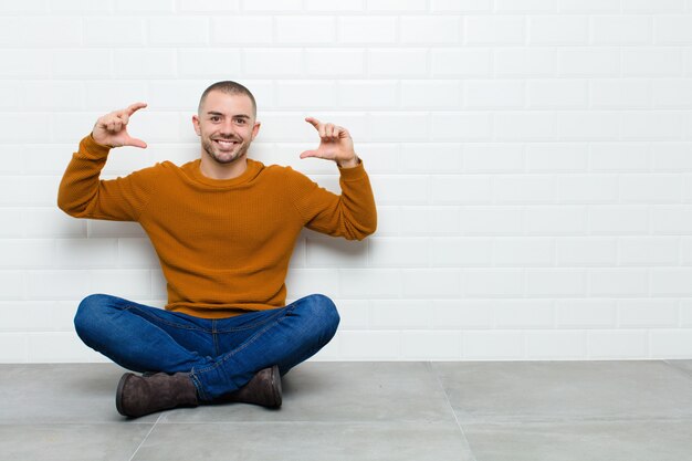 Jeune bel homme encadrant ou décrivant son propre sourire à deux mains, à la recherche de concept de bien-être positif et heureux, assis sur le sol