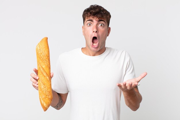 Jeune bel homme étonné choqué et étonné d'une incroyable surprise et tenant une baguette de pain