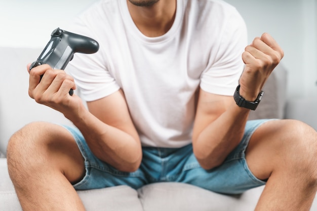 Jeune bel homme excité jouant au jeu vidéo et levant les poings assis sur le canapé à la maison.