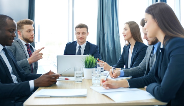 Jeune bel homme faisant des gestes et discutant de quelque chose pendant que ses collègues l'écoutaient assis à la table du bureau