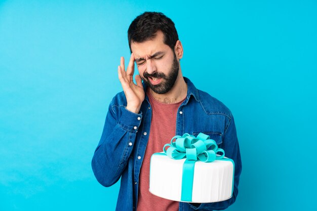 Jeune bel homme avec un gros gâteau sur un mur bleu isolé avec maux de tête