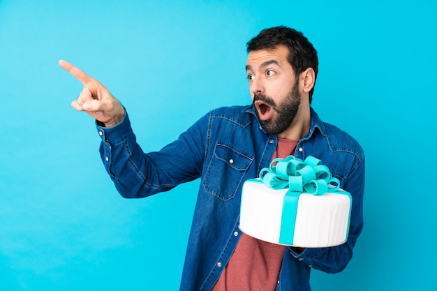 Jeune bel homme avec un gros gâteau sur un mur bleu isolé pointant loin