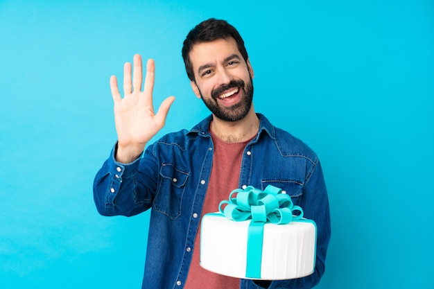 Jeune bel homme avec un gros gâteau saluant avec la main avec une expression heureuse