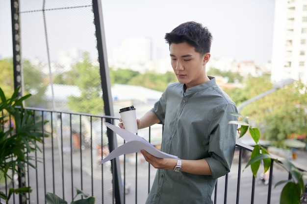 Jeune bel homme heureux assis et lisant un livre, buvant une boisson sur le balcon extérieur