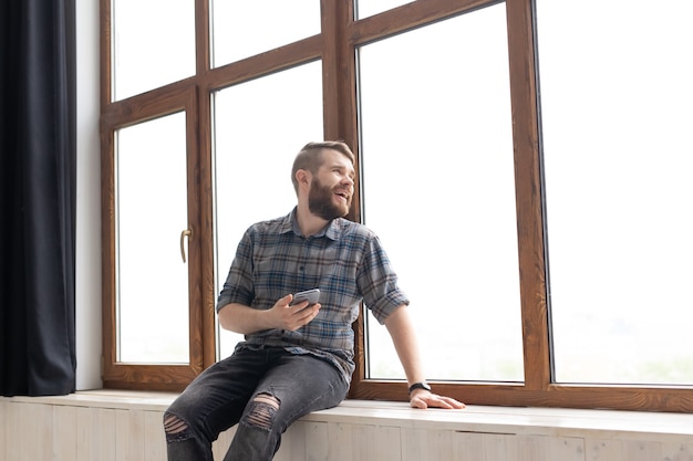 Jeune Bel Homme Hipster Assis Sur Le Rebord De La Fenêtre Près D'une Grande Fenêtre Avec Un Smartphone Dans Son