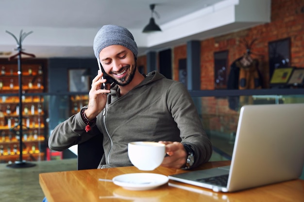 Jeune bel homme hipster avec barbe assis dans un café parlant de téléphone portable tenant une tasse de café et