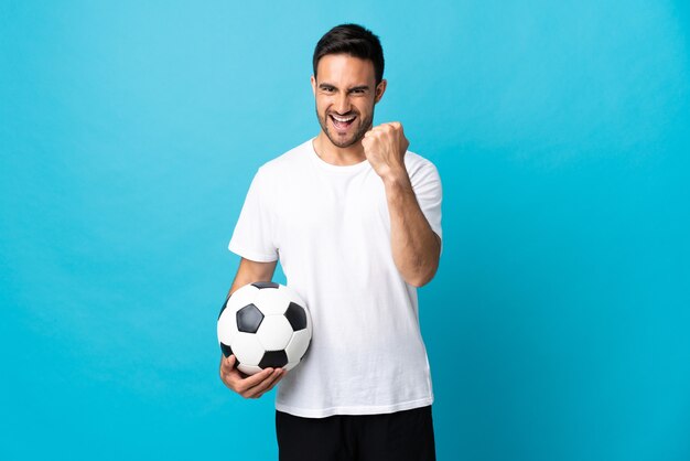 Jeune bel homme isolé sur fond bleu avec ballon de foot célébrant une victoire