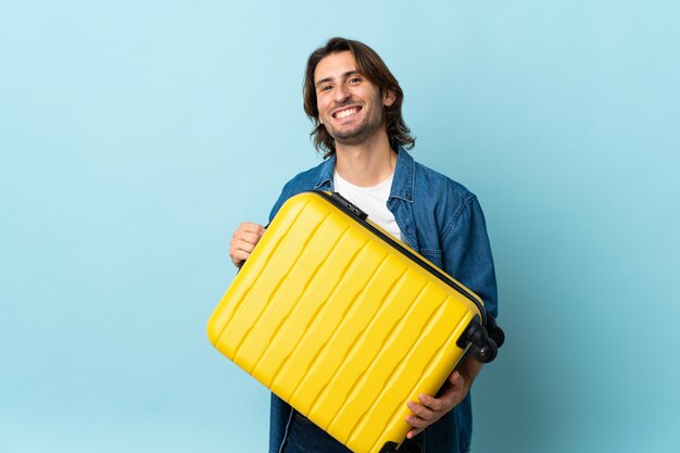 Jeune bel homme isolé sur un mur bleu en vacances avec valise de voyage