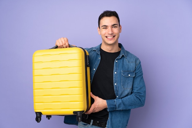 Jeune bel homme isolé sur un mur violet en vacances avec valise de voyage